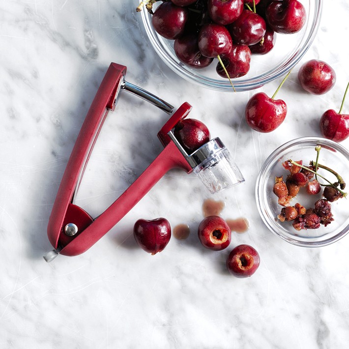Red OXO cherry pitter on a marble counter beside bowls of cherries and pits