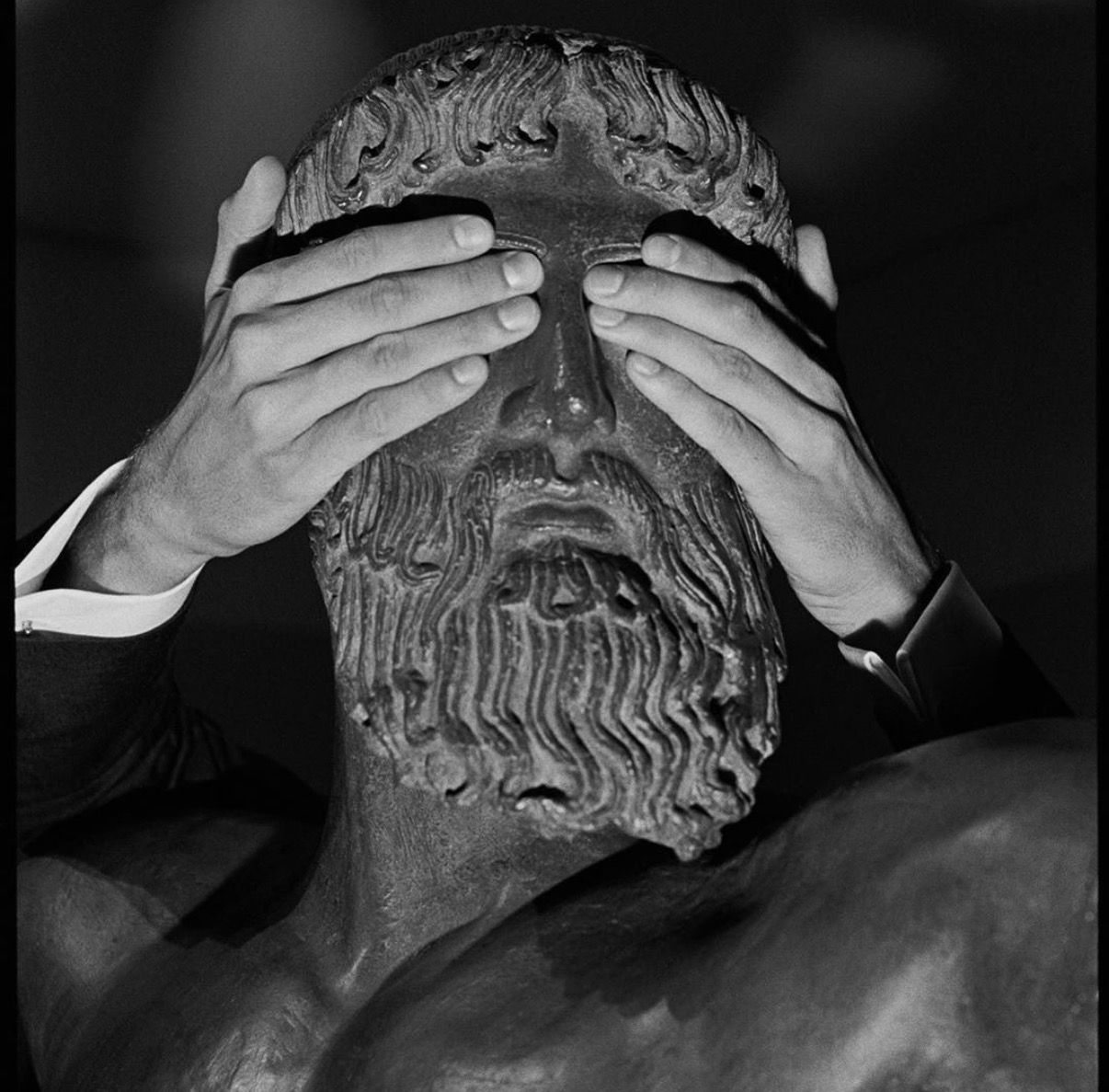 Black-and-white photo of a pair of hands covering the eyes of a bearded bronze statue