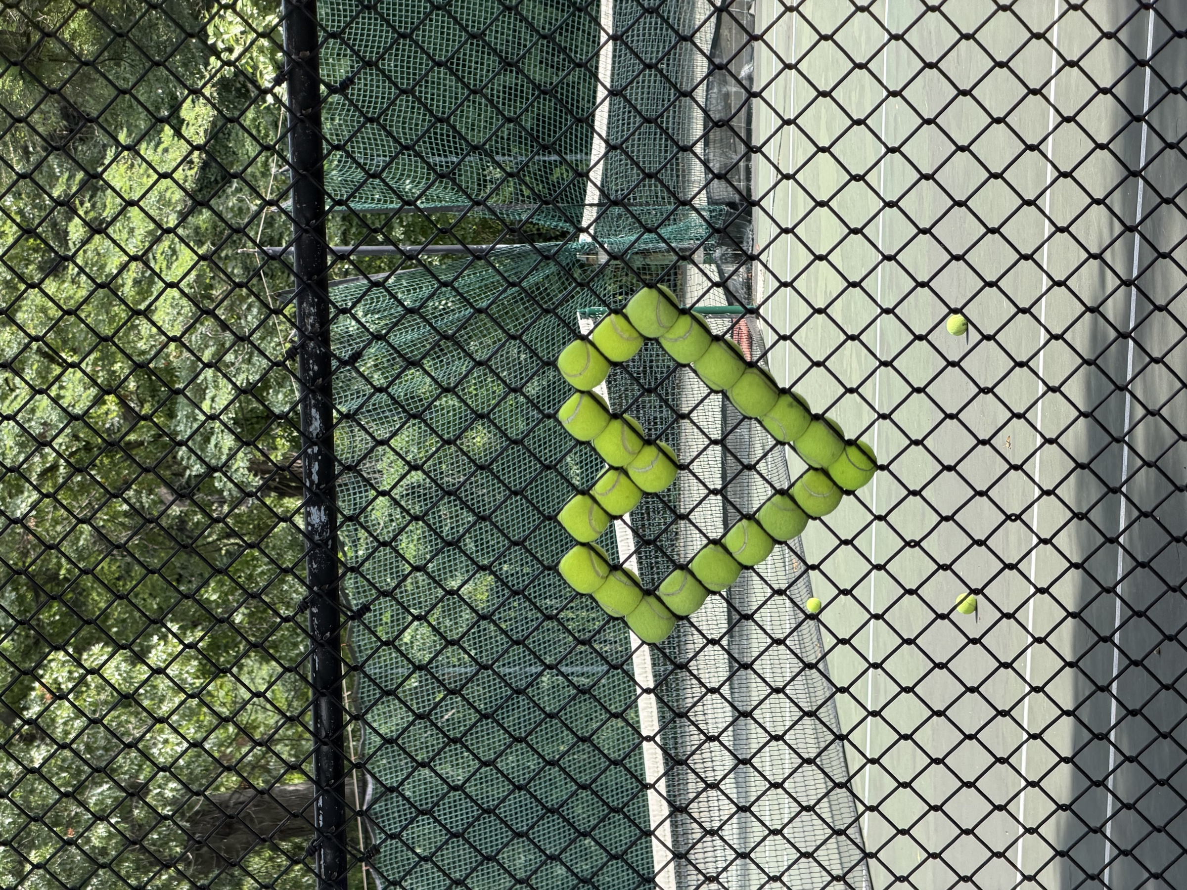 Tennis balls wedged into a chain-link fence in the shape of a heart, in front of a tennis court